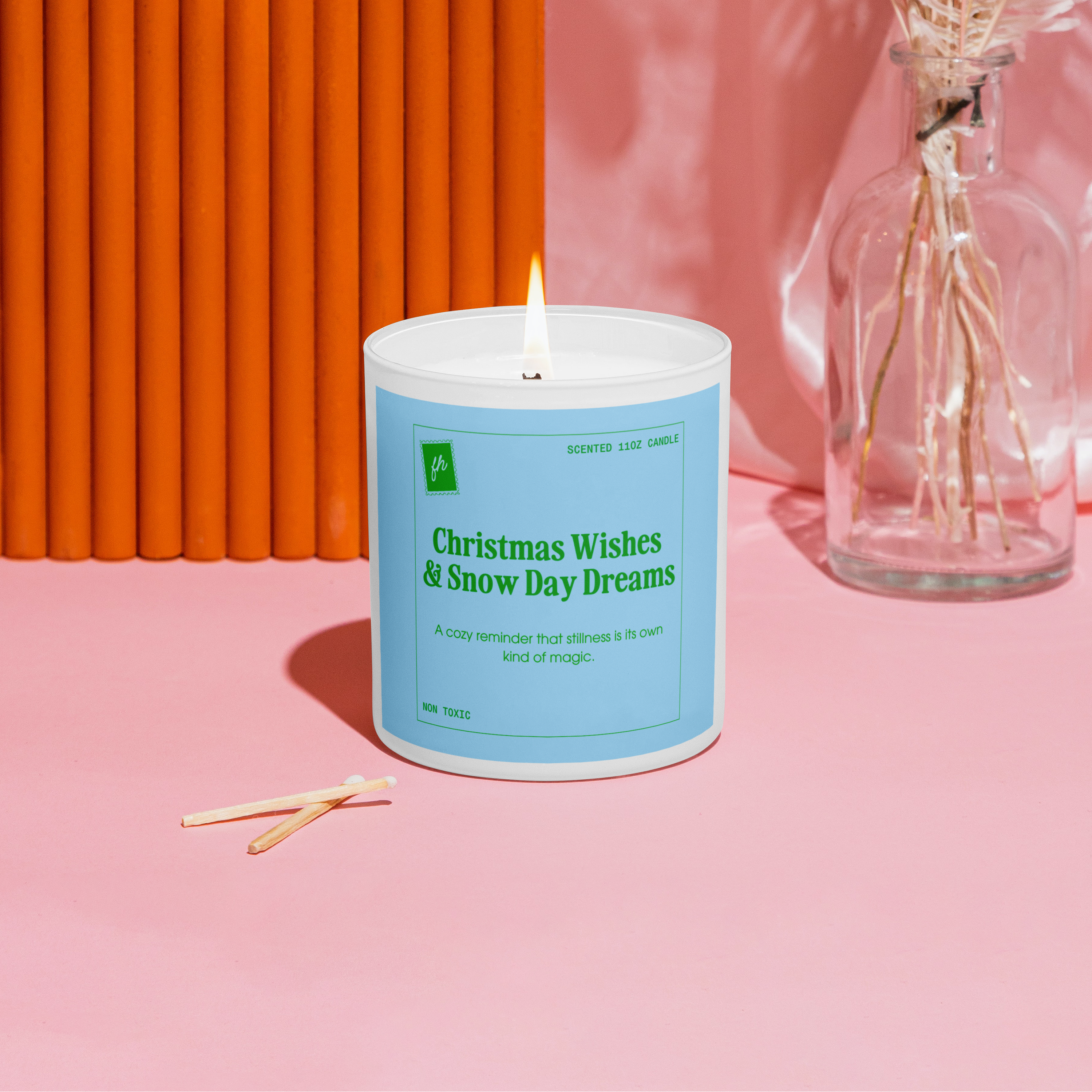 Candle with a blue label on a pink surface with an orange wall and dried flowers in the background
