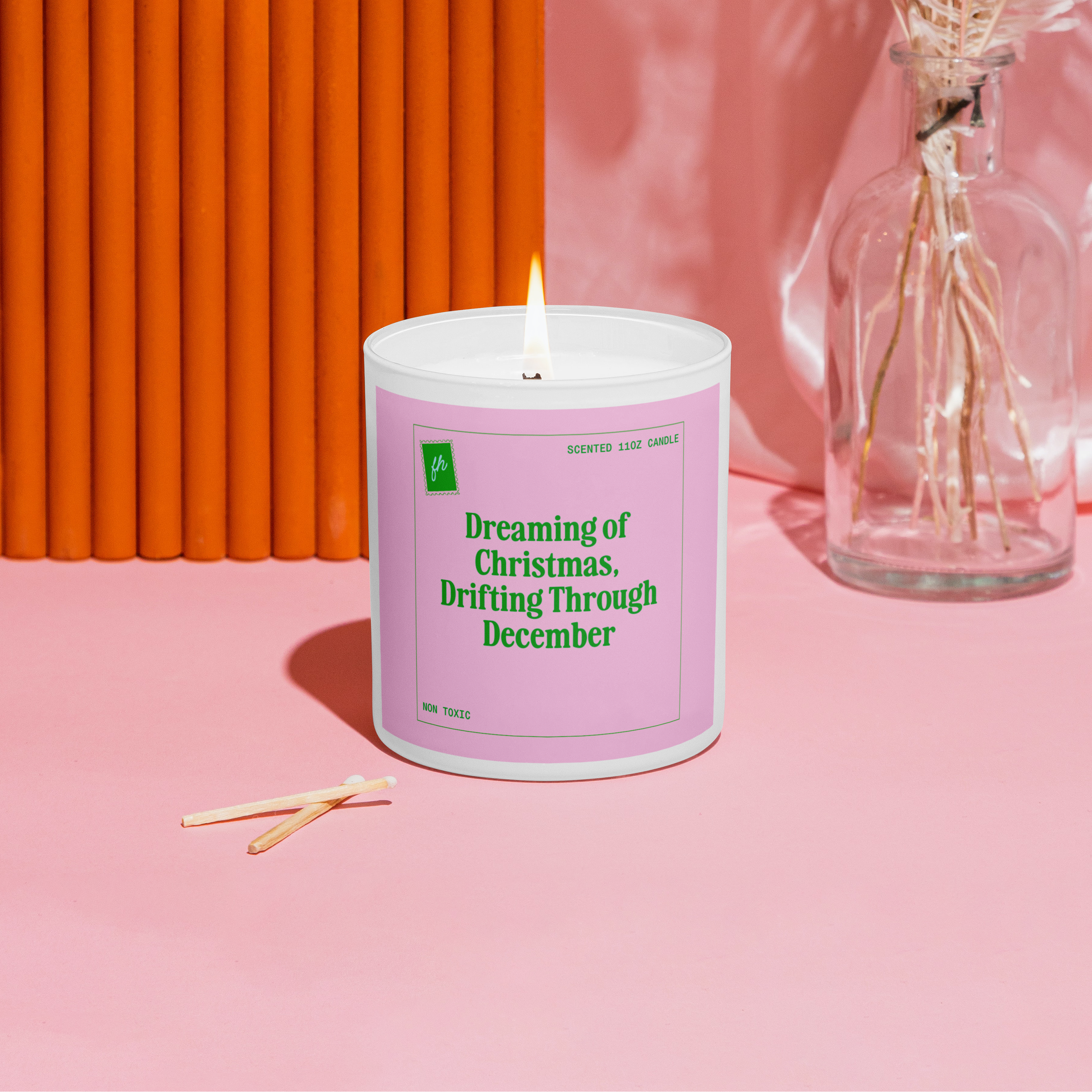 Candle with a pink label on a pink surface with an orange wall and dried flowers in the background