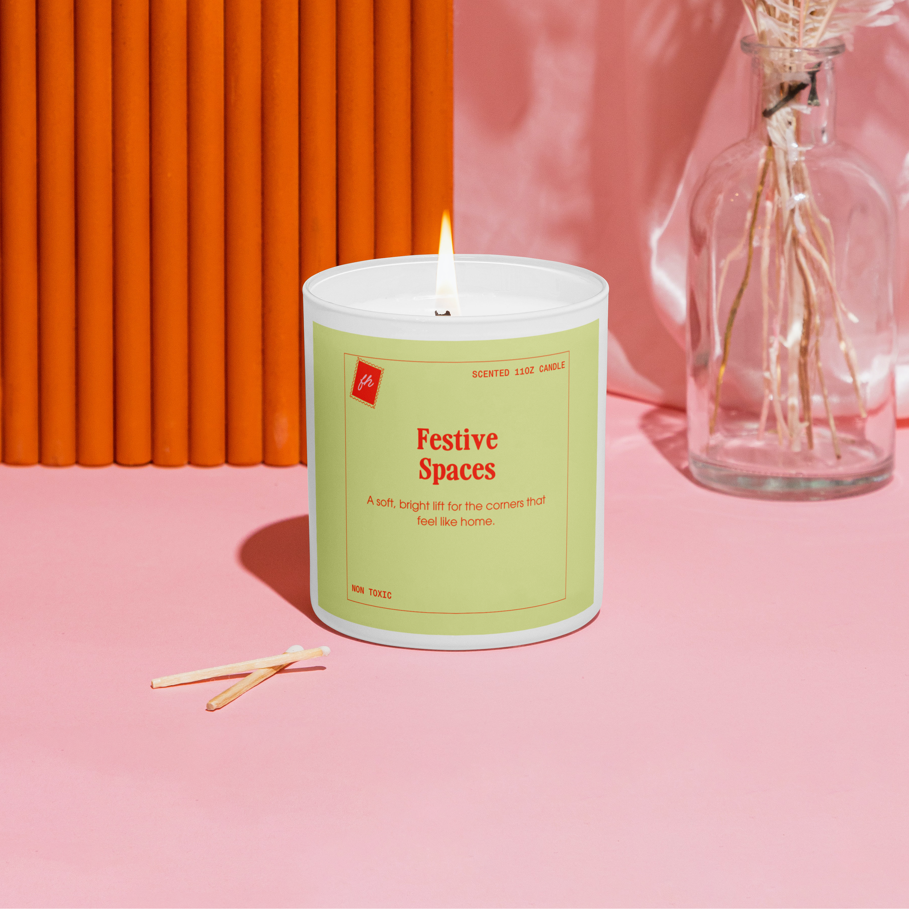 Candle labeled 'Festive Spaces' on a pink surface with an orange wall and dried flowers in the background.