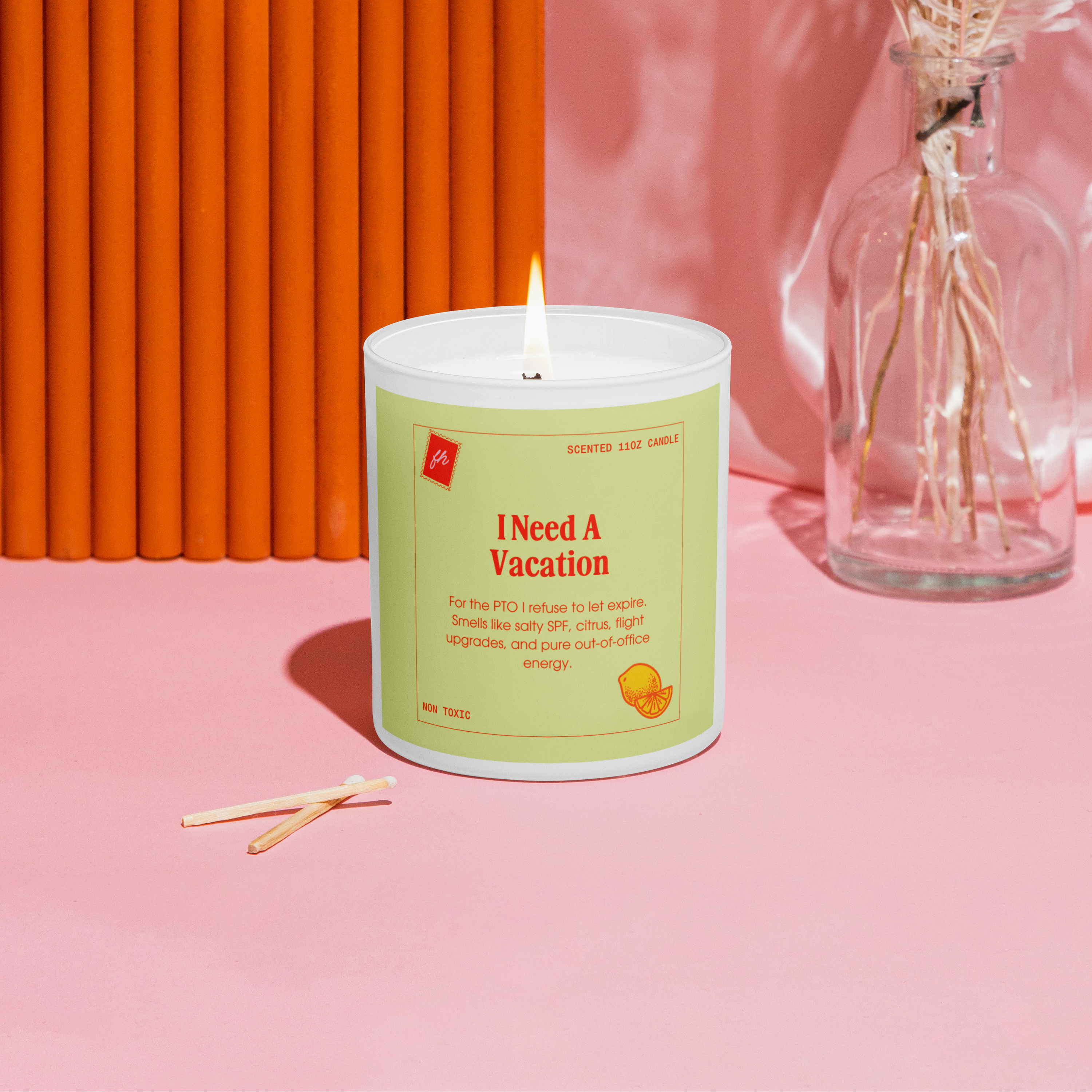 Candle with a label reading 'I Need A Vacation' on a pink surface with an orange wall and dried flowers in the background.