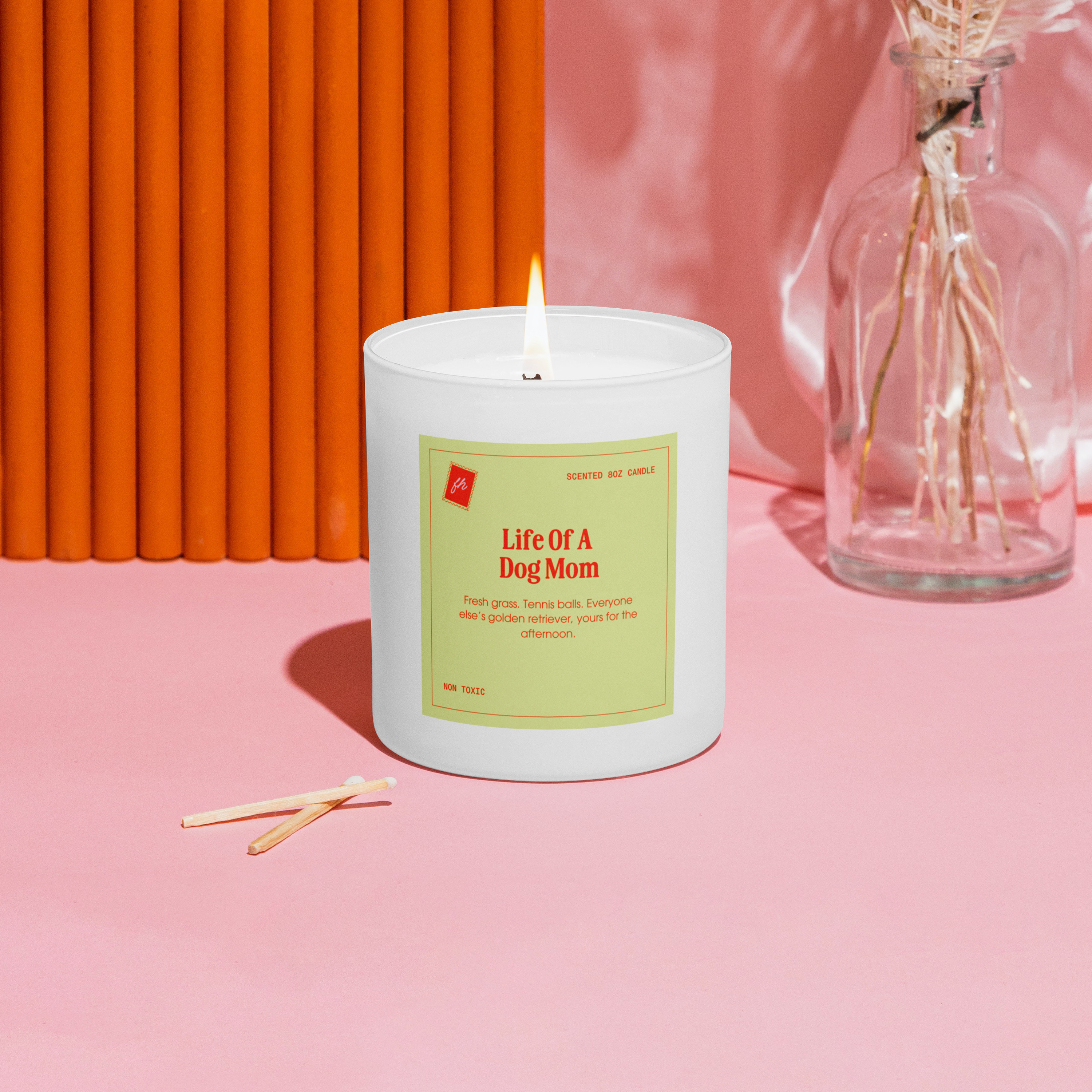 Candle with a label on a pink surface next to a vase with dried flowers against an orange wall.