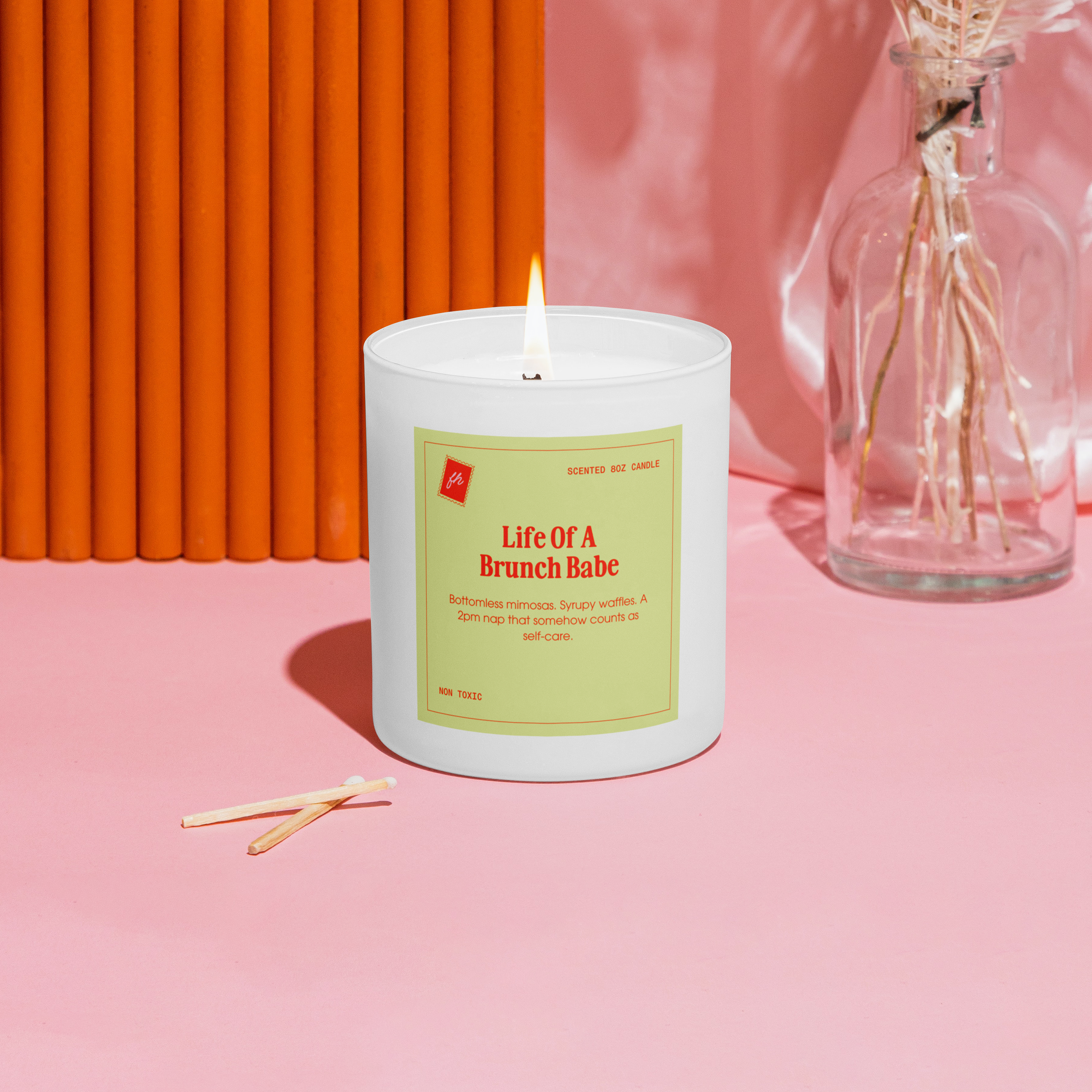 Candle with a label titled 'Life Of A Brunch Babe' on a pink surface with an orange wall and dried flowers in the background.