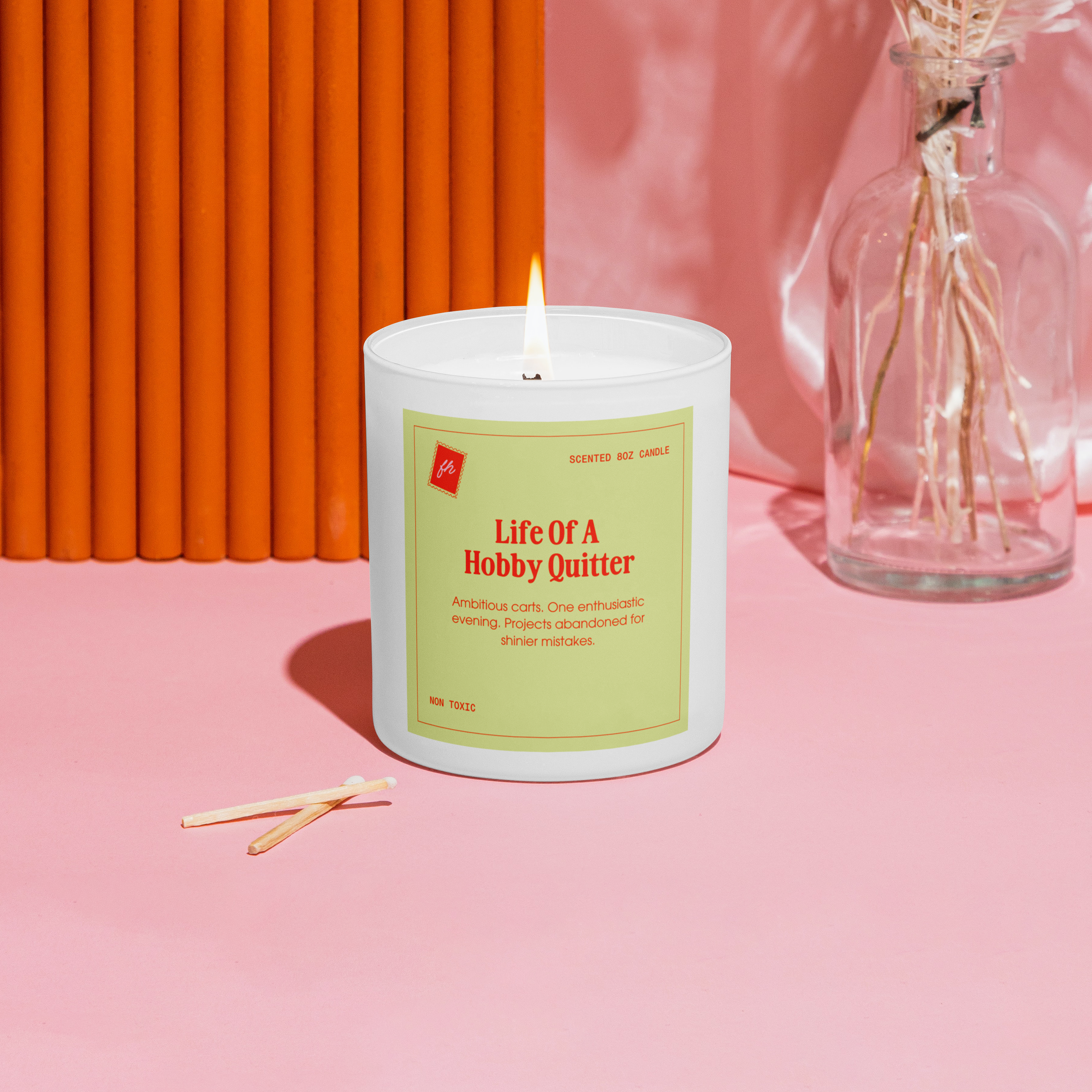 Candle with a label titled 'Life Of A Hobby Quitter' on a pink background with a vase of dried flowers.